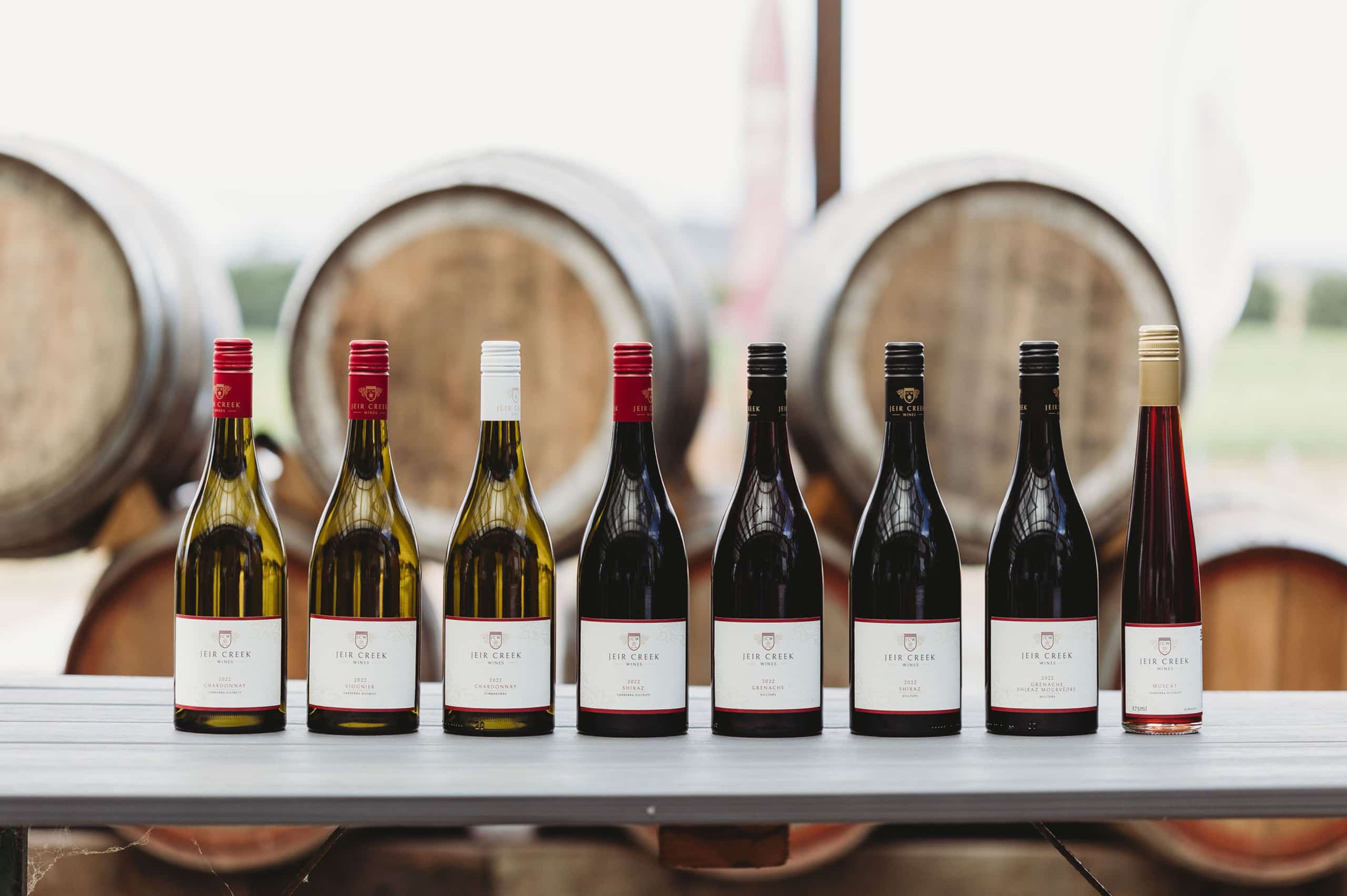 Eight bottles of Jeir Creek Wine sitting side by side on a table in front of wine barrels